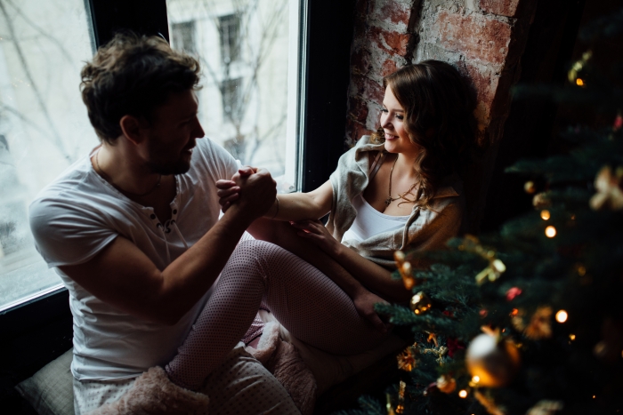 Happy couple of lovers in pajamas sit on the windowsill. Christmas atmosphere and new year gift at home. Young family together. Garland lights in the foreground bokeh.